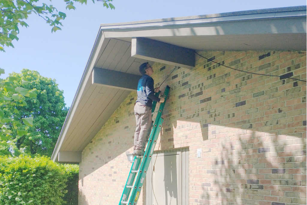 A person looks at the underside of the roof over hang while standing on a ladder leaning against the brick wall of a buliding.