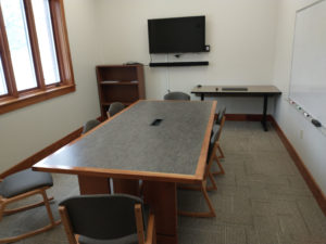 A group meeting room that includes a central table surrounded by seven chairs. Of the three visible walls, one has a large window, another a large television and the last a white board.